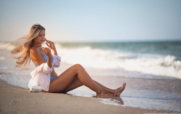 Uma garota com um maiô e uma camisa azulados se senta perto da beira da água e olha para o horizonte