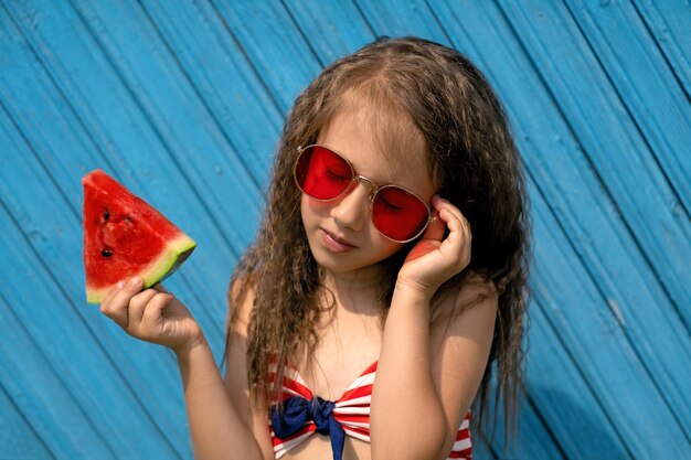 Uma garota com um pedaço de melancia na mão ajusta óculos de sol vermelhos