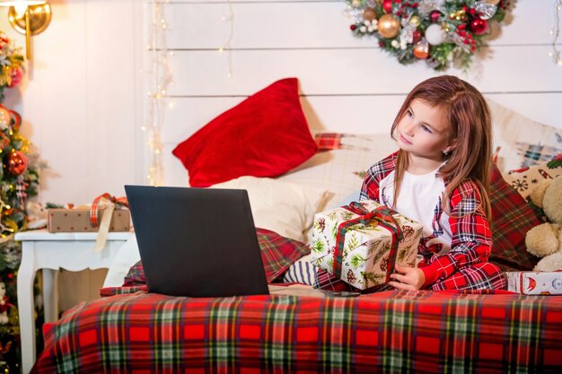 Uma garota com um presente de Natal nas mãos está sentada de pijama em uma cama em frente a um laptop em um quarto decorado.