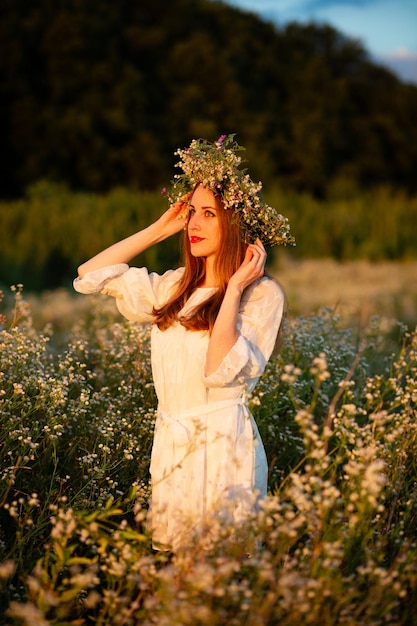 Uma garota com um sorriso fica em um campo com uma coroa na cabeça ao pôr do sol em um vestido branco