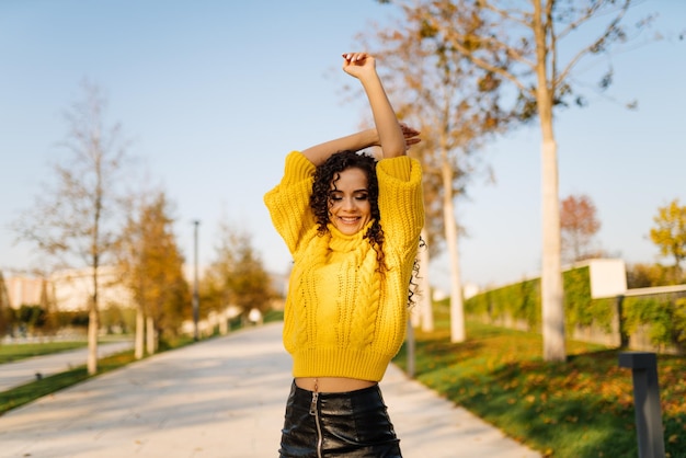 Foto uma garota com um suéter amarelo brilhante dança no caminho do parque de outono levantando as mãos e sorrindo graciosamente