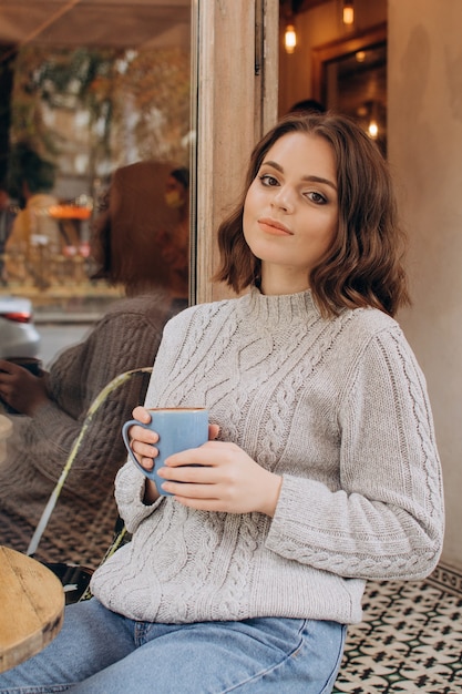 Uma garota com um suéter cinza se senta a uma mesa em um café, bebe chocolate e aprecia o aroma