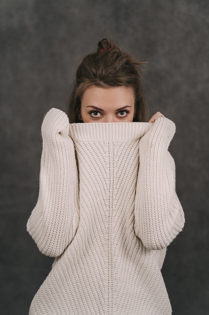 Uma garota com um suéter claro em uma parede cinza. A garota esconde o rosto atrás do suéter. A garota só pode ver seus olhos.