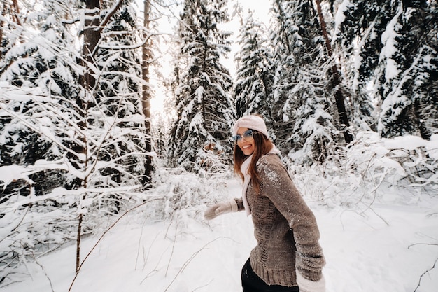 Uma garota com um suéter e óculos caminha na floresta coberta de neve no inverno.