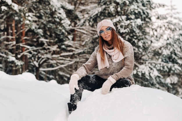 Uma garota com um suéter e óculos no inverno senta-se sobre um fundo coberto de neve na floresta.