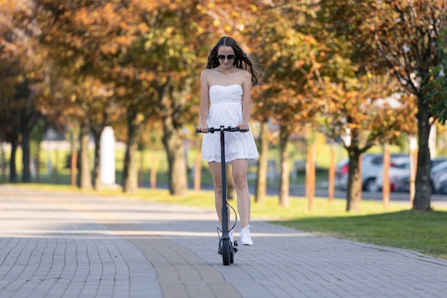 Uma garota com um vestido de verão anda de scooter elétrica na calçada