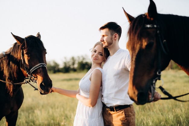 Uma garota com um vestido de verão branco e um cara com uma camisa branca em uma caminhada com cavalos marrons na aldeia