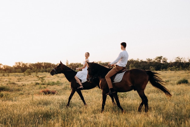 Uma garota com um vestido de verão branco e um cara com uma camisa branca em uma caminhada com cavalos marrons na aldeia