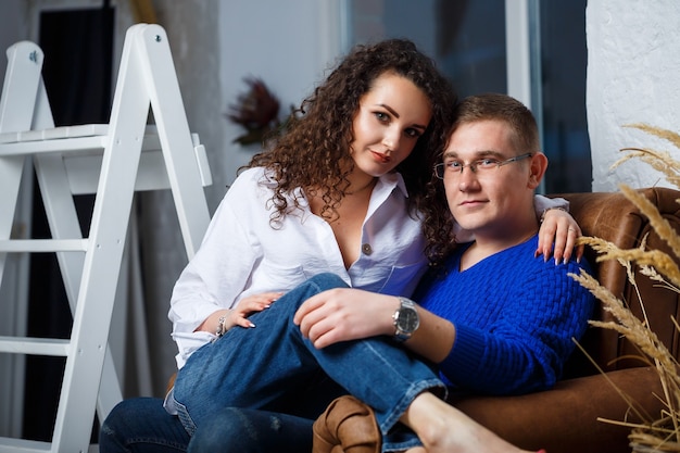 Foto uma garota com uma camisa branca e um cara com um swirere azul estão sentados em uma poltrona e se abraçando, beijando, ternura, amor