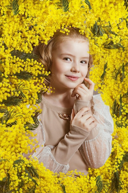 Uma garota com uma jaqueta bege está cercada por flores de mimosa