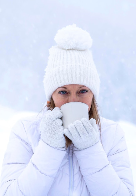 Uma garota com uma jaqueta branca e um chapéu branco está segurando uma xícara de chá nas mãos durante uma nevasca no contexto de lindas nevascas
