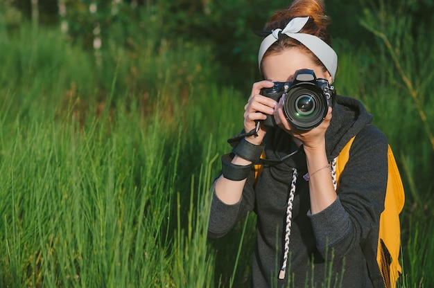 Uma garota com uma jaqueta cinza e uma mochila amarela segura uma câmera fotográfica profissional nas mãos. no contexto da natureza verde e da floresta.