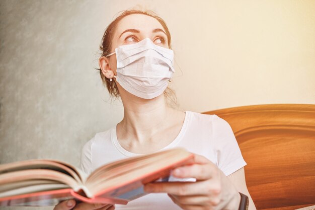 Uma garota com uma máscara médica está em quarentena em casa, lendo um livro e olhando pela janela.