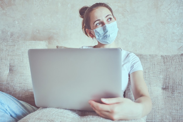 Uma garota com uma máscara médica está em quarentena em casa, segurando um laptop e olha pela janela.