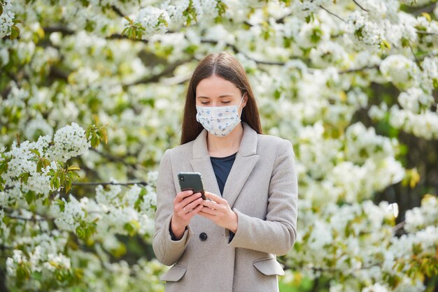 Uma garota com uma máscara médica para evitar a propagação do coronavírus lê as notícias em um smartphone. Uma mulher com uma máscara facial contra o COVID-19 mantém uma distância social no jardim entre árvores floridas.