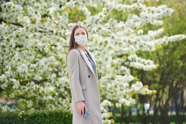 Uma garota com uma máscara médica para evitar a propagação do coronavírus segura um smartphone no parque. Uma mulher com uma máscara facial contra o COVID-19 mantém uma distância social no jardim entre árvores floridas.