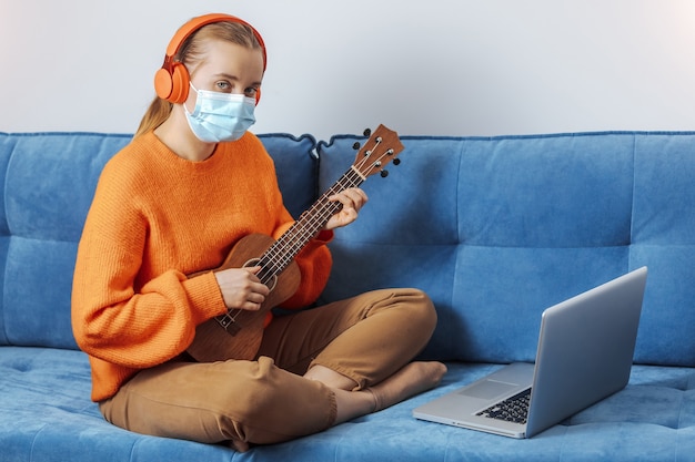 Uma garota com uma máscara médica se senta no sofá em casa e aprende a tocar ukulele remotamente