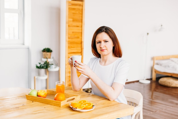 Uma garota de aparência europeia senta-se à mesa com uma camiseta branca, laranjas para fazer smoothies e sucos, os benefícios das vitaminas, manhã