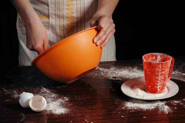 Uma garota de avental em uma cozinha escura amassa a massa em uma xícara de laranja.