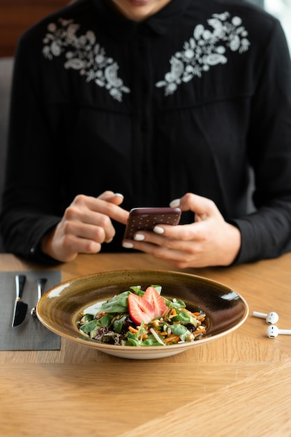 Uma garota de blusa preta fotografa comida em um restaurante com um smartphone. Salada de legumes decorada com morangos frescos. Profundidade de campo rasa, fundo desfocado.