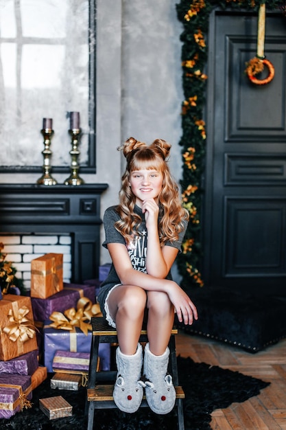 Uma garota de cabelo branco está sentada no chão perto de uma árvore de Natal e uma caixa de presentes