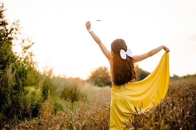 Uma garota de cabelo comprido e um laço branco fica em uma saia amarela em um campo com espigas