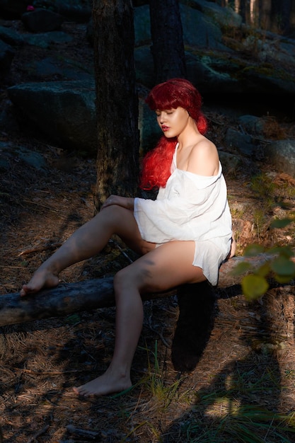 Uma garota de cabelo vermelho e uma camisa branca com uma rosa nas mãos está descansando em uma floresta fabulosa