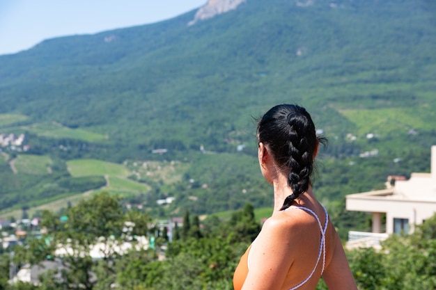 Uma garota de cabelos escuros olha para o lado em um maiô vermelho em um vale de montanha com vinhedos. Vista traseira.