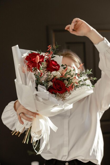 Uma garota de camisa branca segura um buquê de flores contra uma parede preta Lótus de eucalipto rosa vermelha