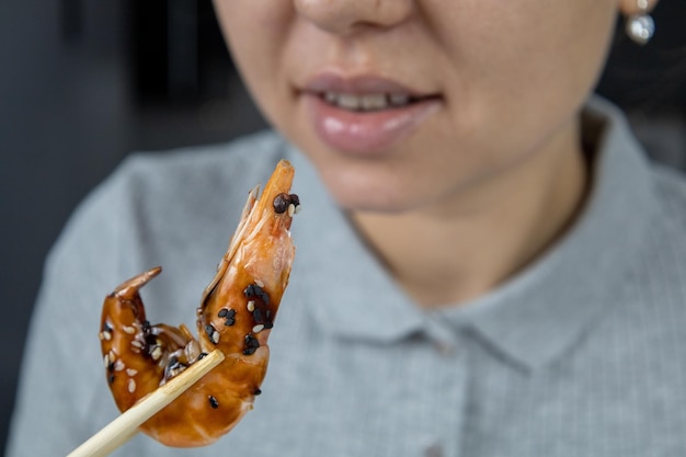 Uma garota de camisa cinza na cozinha segura um camarão rei em molho unagi e sementes de gergelim com pauzinhos Conceito de comida asiática