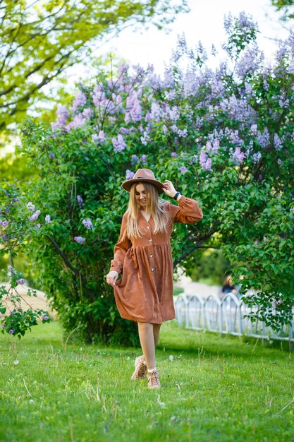 Uma garota de chapéu e vestido marrom caminha pelo parque, ela tem uma aparência europeia e um sorriso no rosto. Mulher jovem e atraente caminhando em um dia ensolarado