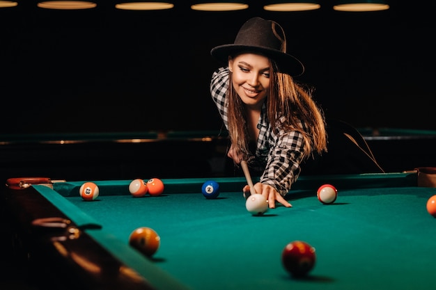 Uma garota de chapéu em um clube de bilhar com um taco nas mãos acerta uma bola. Jogando bilhar.