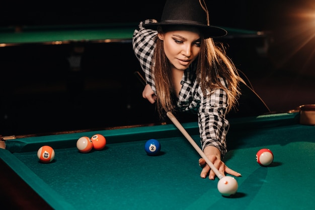 Uma garota de chapéu em um clube de bilhar com um taco nas mãos acerta uma bola. Jogando bilhar.