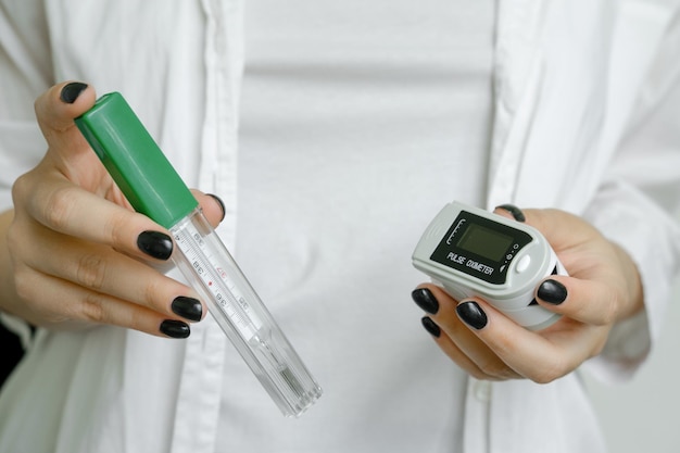 Uma garota de jaleco branco segura um termômetro para medir a temperatura corporal e um oxímetro de pulso covid