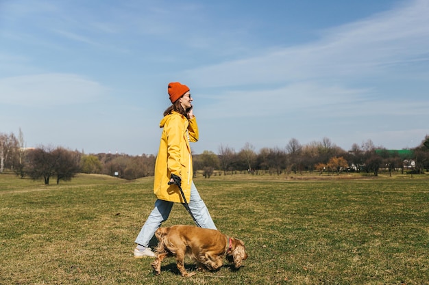 uma garota de jaqueta amarela caminha com um cachorro spaniel no parque e fala ao telefone