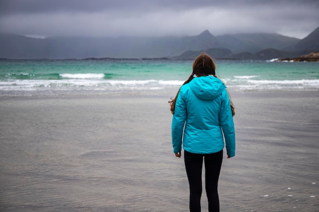 uma garota de jaqueta azul admira a paisagem sombria em uma praia nas ilhas lofoten na noruega