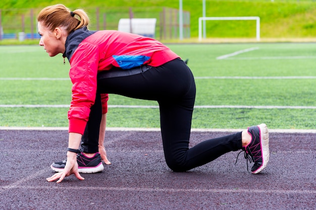 Uma garota de legging preta e um blazer rosa preparado para correr em uma esteira em uma arena ao ar livre e está em uma pose de corredores