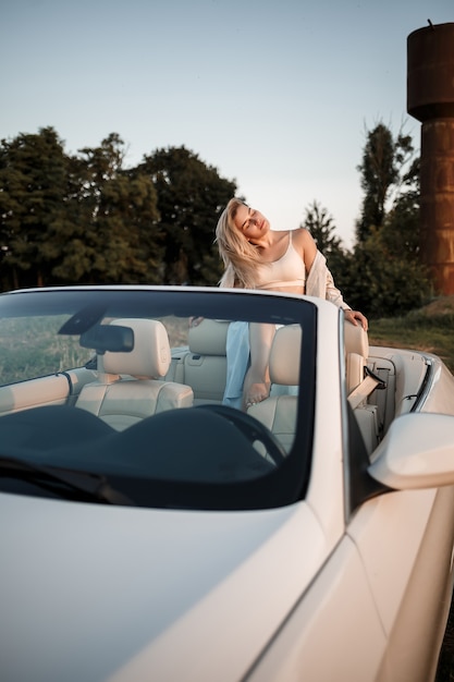 Foto uma garota de luxo glamourosa com cabelo loiro sorrindo enquanto está sentado em um conversível branco. jovem mulher de sucesso sentada em seu carro branco