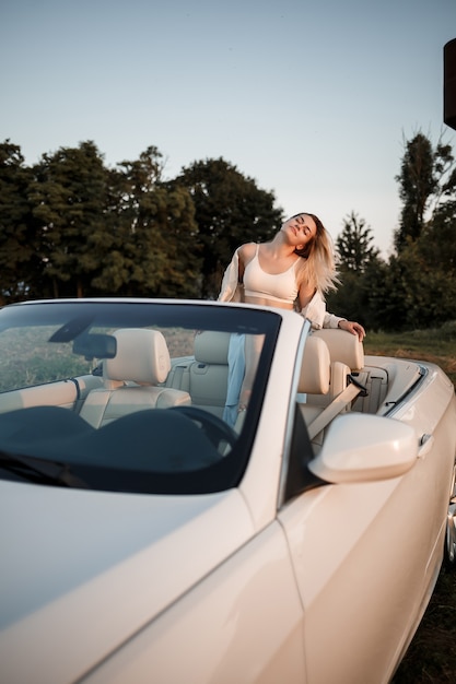 Uma garota de luxo glamourosa com cabelo loiro sorrindo enquanto está sentado em um conversível branco. jovem mulher de sucesso sentada em seu carro branco