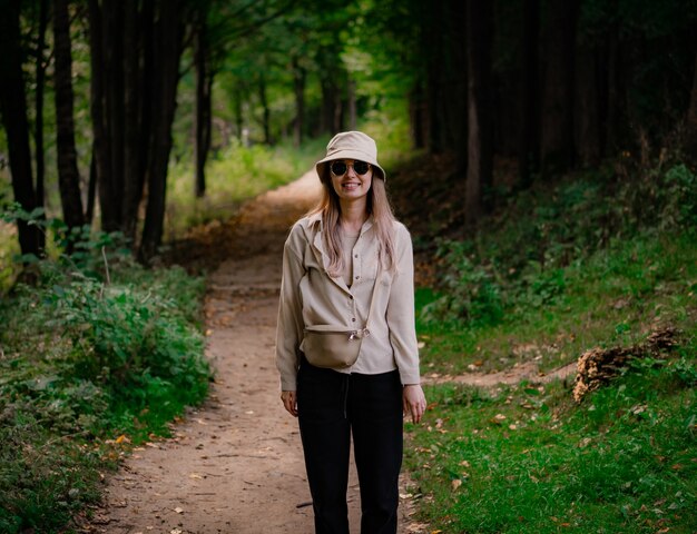 Uma garota de óculos escuros e chapéu bege está na floresta e sorri