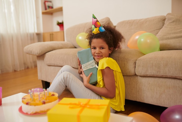 Uma garota de pele escura com um chapéu de aniversário sentada no chão e segurando um presente