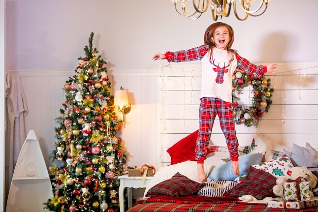 Uma garota de pijama vermelho com um cervo pula em uma cama ao lado de uma árvore de Natal.