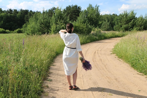 Uma garota de roupas brancas caminha por uma estrada rural com um buquê de flores silvestres