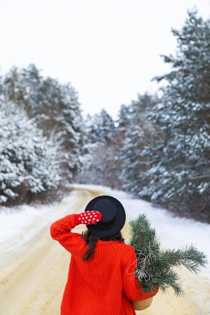 Uma garota de suéter vermelho e chapéu fica no meio de uma estrada de neve em uma floresta com galhos de pinheiro Viagem de fim de semana