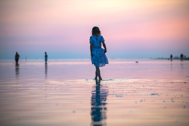 Uma garota de vestido à beira-mar ao pôr do sol caminha sobre a água