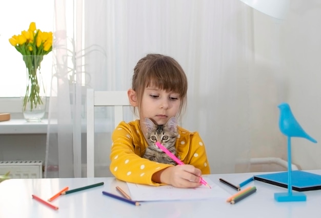 Uma garota de vestido amarelo desenha em seu quarto em sua mesa com lápis de cor petscats ao lado dela em casa O conceito de criatividade e desenvolvimento infantil Foco seletivo