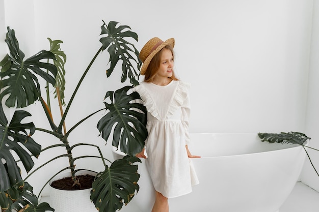 uma garota de vestido branco e um chapéu de palha em uma sala com plantas e um banheiro