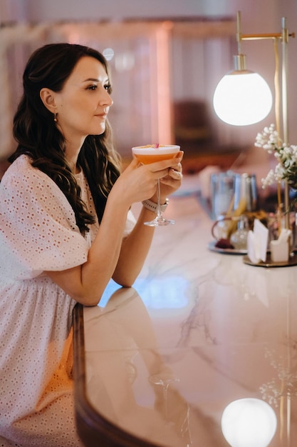 Uma garota de vestido branco está sentada no bar de um café e bebendo um coquetel