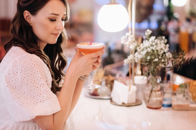 Uma garota de vestido branco está sentada no bar de um café e bebendo um coquetel