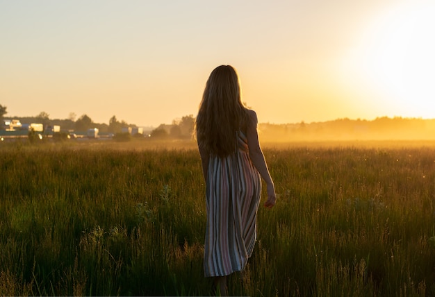 Uma garota de vestido caminha por um campo com neblina ao pôr do sol em direção ao sol